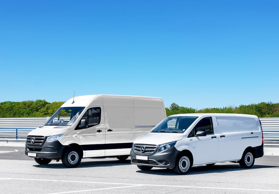 Two white Mercedes-Benz vans are parked side by side on a paved road with clear blue sky overhead and a backdrop of green trees and a metal barrier. The larger van on the left features a high roof and