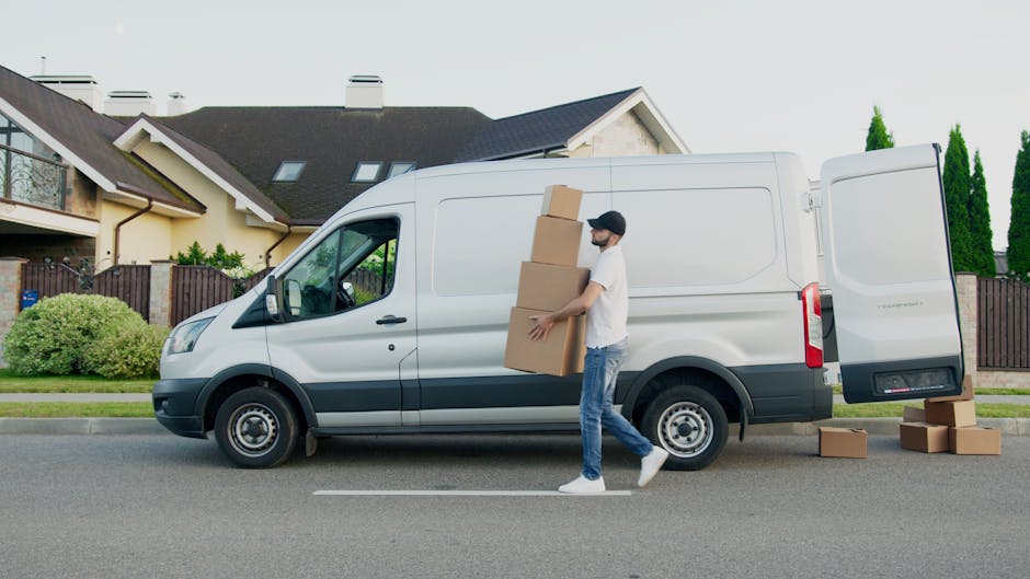 Two white Mercedes-Benz vans are parked side by side on a paved road with clear blue sky overhead and a backdrop of green trees and a metal barrier. The larger van on the left features a high roof and