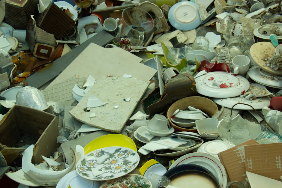A large pile of broken and discarded household items, including shattered ceramic plates with floral patterns, broken cups, bowls, and glass fragments, mixed with packaging materials such as cardboard boxes, foam, and plastic wrapping. The debris is situated outdoors on a pavement or loading area near a property entrance, with some objects overlapping and scattered unevenly. Visible in the background are additional broken ceramics, damaged kitchenware, and miscellaneous broken household items. The scene appears to be part of a rubbish clearance or disposal process related to home relocation or furniture transport, illustrating the type of waste collected during a comprehensive removal service. The lighting is natural daylight, highlighting the variety of materials and the disorganized nature of the waste, managed or prepared for collection by [COMPANY_NAME], a professional removals service specializing in house clearances and moving logistics.