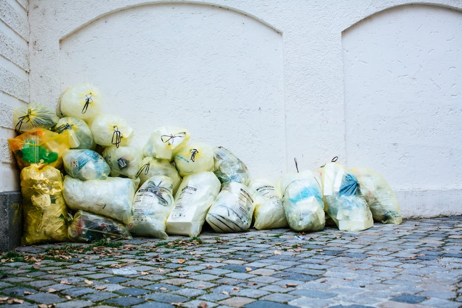 A collection of yellow and white garbage bags filled with household waste, placed on a cobblestone pavement next to a white exterior wall with a brick corner visible to the left. Some bags appear to contain paper or plastic packaging materials, and they are tied with black drawstrings. The bags are stacked and lined up along the wall, likely awaiting disposal or collection. The setting suggests a scene during or after a house clearance or packing process as part of a home relocation or move. This image reflects the type of bulky waste pickup service that Man With a Van Putney offers in Putney, focusing on safe disposal of household waste without fines, aligned with the page's topic of bulky waste removal and furniture transport in the context of removals.
