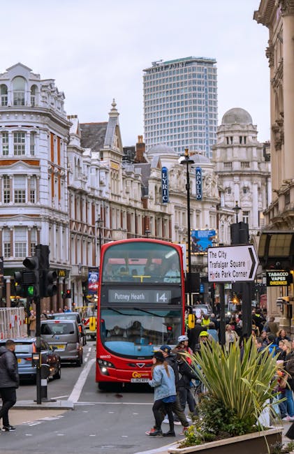 A busy street scene in Putney showing a red double-decker bus with route number 14 and destination Putney Heath approaching a pedestrian crossing. Surrounding the bus are various vehicles, including cars waiting at a traffic light, and pedestrians crossing the road, some carrying bags or briefcases. Buildings with ornate Victorian architecture line the street, with several shopfronts and signs visible, including a McDonald's and a Tabacco shop. In the background, a modern high-rise building rises behind historic structures, and a street sign indicates directions to The City, Trafalgar Square, and Hyde Park Corner. The scene is set during daytime with overcast sky, and greenery such as a large potted plant is visible in the foreground, near the pavement. This image relates to urban transport and street activity in Putney, in the context of home relocation and moving services, exemplifying typical environments where Man With a Van Putney offers removals and furniture transport solutions.