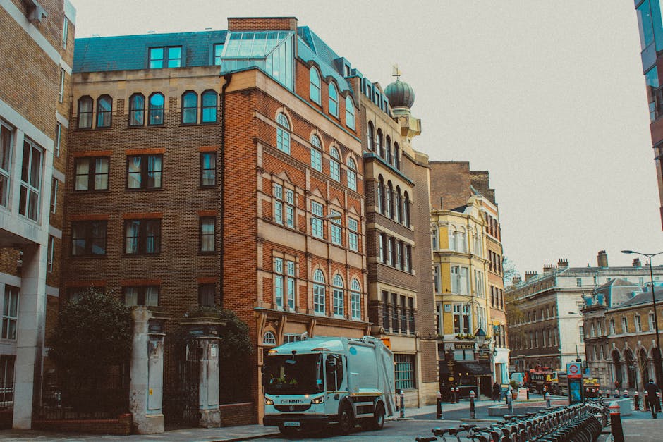 A white moving van is parked on a city street in front of tall, historic brick and stone buildings with large windows, some of which have decorative arches. The van is positioned near a sidewalk, and there are several large cardboard boxes, wrapped with plastic and packed with packing materials, placed on the pavement next to it, indicating a home relocation or furniture transport process. The loading area of the van is open, and a person is visible lifting or carrying an item towards the vehicle. Surrounding the scene are other urban elements such as a bicycle rack, street lamps, and additional buildings with classical architectural features, suggesting an ongoing move or packing activity in an urban environment consistent with house removals services offered by Man With a Van Putney, aligned with the costs and quotes guidance on the Putney removals cost page.