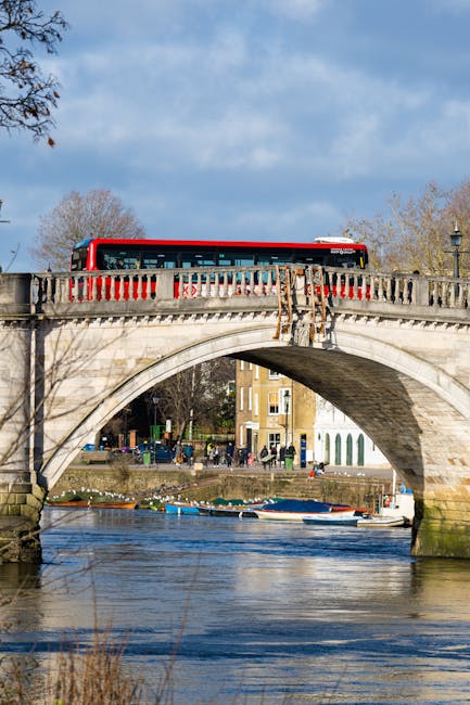 A scenic view of the River Thames near Putney Bridge during daytime, with calm water reflecting the blue sky and scattered white clouds above. Several small boats, made of wood and painted in green an