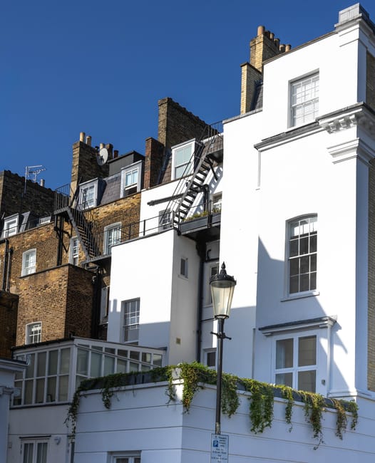 The image depicts the exterior of a multi-story residential building on a Putney estate, with white-painted walls contrasting against neighboring brown brick structures. Fire escape stairs made of black metal are attached to the back of the building, extending from the higher floors down to the ground, and installed alongside several windows. A tall, traditional black street lamp is positioned in front of the building near the pavement, which features a small white parking sign. On a lower level, a balcony area with glass enclosures and lush green plants draping over the edge is visible, indicating outdoor space for residents. The scene is lit by natural daylight, with clear blue skies overhead, highlighting the building's architecture and surroundings. This setting exemplifies the urban environment where house removals and furniture transport services by Man With a Van Putney are often required, especially when navigating staircases and parking challenges on busy estates.