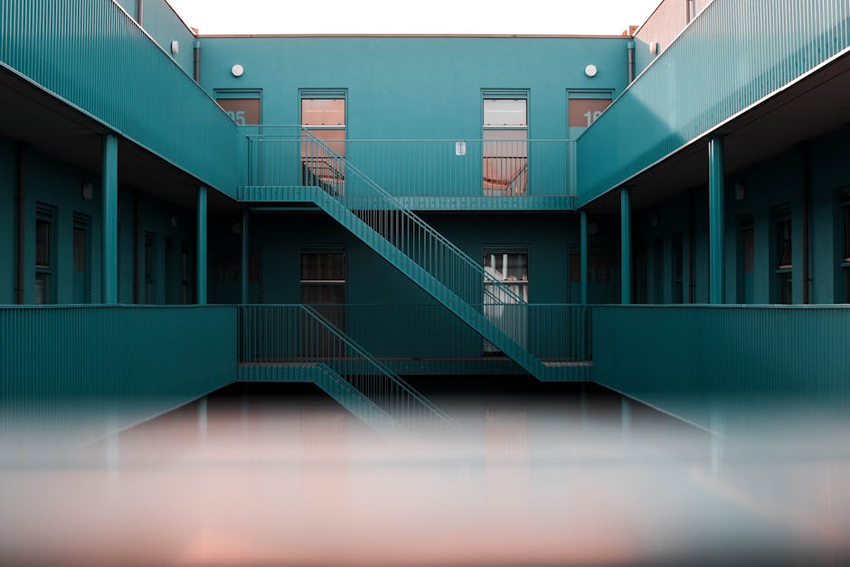 A multi-storey residential building with exterior staircases made of metal, painted in teal, connecting the ground and upper floors, with open walkways and doorways visible on each level. The building features a modern, utilitarian design with railings along the walkways and staircases, and a flat roof with numbered apartment entrances seen through the open doorways. The scene is captured during daylight with natural lighting, providing a clear view of the structural layout designed for access and egress. This setup highlights typical architectural elements that may pose challenges for home relocations, such as moving furniture or boxes through staircases and narrow corridors, and illustrates the importance of professional moving services like those offered by Man With a Van Putney, especially when navigating such staircase and parking constraints on Putney estates.