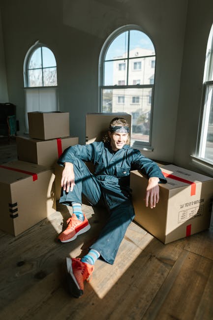 A young man with long hair, wearing a denim jacket, dark pants, blue socks, and bright orange sneakers, is sitting on a wooden floor inside a well-lit room with large arched windows. He is leaning against a large cardboard box secured with red packing tape, with several other unopened boxes of various sizes stacked nearby. The sunlight streaming through the windows illuminates the scene, highlighting the boxes and the man’s relaxed posture. The room appears to be during a home relocation process, with the boxes ready for moving, and the background shows a view of neighboring buildings through the windows. The environment suggests an interior space prepared for furniture transport and packing as part of a house removal service, with [COMPANY_NAME] performing the moving logistics, including loading and packing assistance.