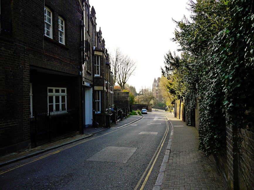 A quiet residential street in Putney with a narrow pavement on the right side, bordered by a tall brick wall and dense green foliage. On the left, there are multi-storey brick houses with bay windows, some with small front gardens enclosed by black metal railings. The asphalt road has double yellow lines along the curb and a few parked cars visible in the distance. In the foreground, an area near the entrance of a house shows a pile of packed cardboard boxes, wrapped furniture, and a moving blanket, indicating recent packing for a house relocation. Man With a Van Putney is involved in an ongoing furniture transport or packing and moving process, as evidenced by the arrangement of items prepared for loading onto a van, which is partially visible out of frame. The scene is illuminated by natural daylight, with some shadows cast by the trees and buildings, suggesting a morning or late afternoon setting.