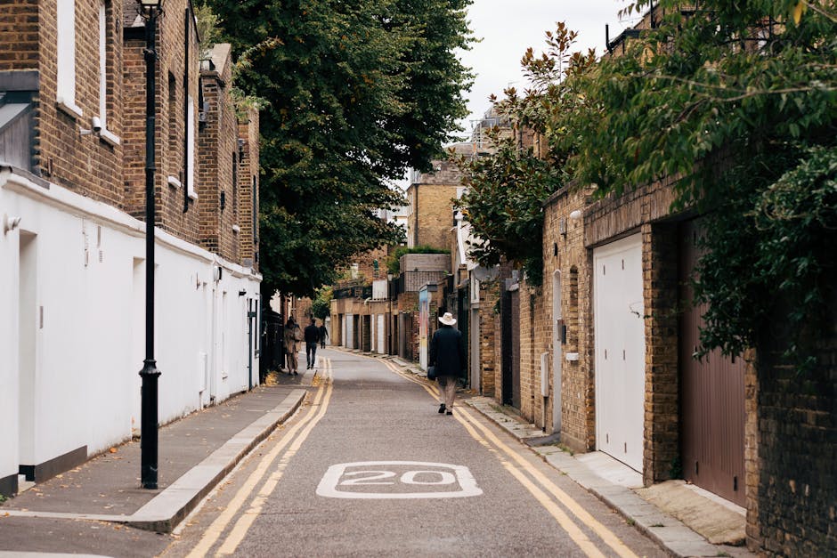A straight street scene showing a residential and commercial area in Putney, with parked cars lining both sides of the road and pedestrians walking along the pavement. The street is paved with asphalt and has white dashed lane markings in the centre. On the left side, there is a white building with a black sign, along with large flower pots containing colorful flowers placed on the pavement. On the right side, a row of buildings with shopfronts and storefront signage is visible, including a pharmacy. Several vehicles, such as a black hatchback, a silver sedan, and a dark blue car, are parked along the street on both sides. The street is flanked by trees with lush green foliage, providing partial shade to the area. This scene exemplifies typical urban street activity suitable for home relocation and furniture transport services, with the environment prepared for loading or unloading in the context of house removals, as handled by Man With a Van Putney.