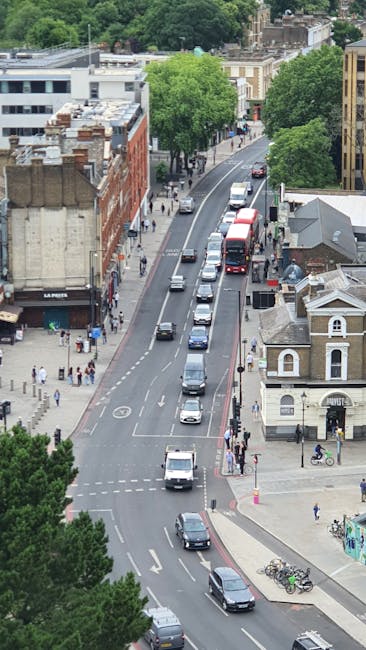 An aerial view of a busy urban street in Putney, with parked and moving vehicles including cars and a double-decker bus on a curved road lined with trees and mixed architecture, such as residential houses and commercial buildings. Sidewalks are populated with pedestrians, some waiting or walking, and bicycles are parked near a corner building. The scene captures daytime lighting with green trees providing contrast to the grey and brick facades. The image reflects an active area typical for relocation logistics, where furniture transport and home removals might occur with careful planning within city streets, as represented in the context of house removals services by Man With a Van Putney.