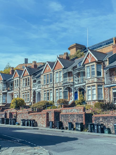 Several Victorian-style terraced houses with decorative white ironwork and bay windows line the street in Putney, London, under a bright blue sky with some scattered clouds. The houses are set on a slightly elevated area behind a brick retaining wall, which features a row of black wheelie bins and waste containers along the pavement. In the foreground, a man with a van belonging to Man With a Van Putney is involved in a home relocation process, with the open back of the van visible, presumably being loaded with furniture, boxes, and packing materials prepared for a residential move as part of the house removals service. The scene captures the typical urban environment of a suburban street, emphasizing the logistics involved in furniture transport and packing for a smooth moving experience.