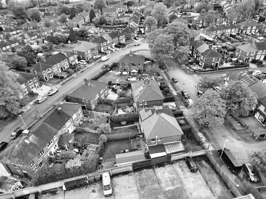 An aerial view of a busy urban street in Putney, with parked and moving vehicles including cars and a double-decker bus on a curved road lined with trees and mixed architecture, such as residential houses and commercial buildings. Sidewalks are populated with pedestrians, some waiting or walking, and bicycles are parked near a corner building. The scene captures daytime lighting with green trees providing contrast to the grey and brick facades. The image reflects an active area typical for relocation logistics, where furniture transport and home removals might occur with careful planning within city streets, as represented in the context of house removals services by Man With a Van Putney.