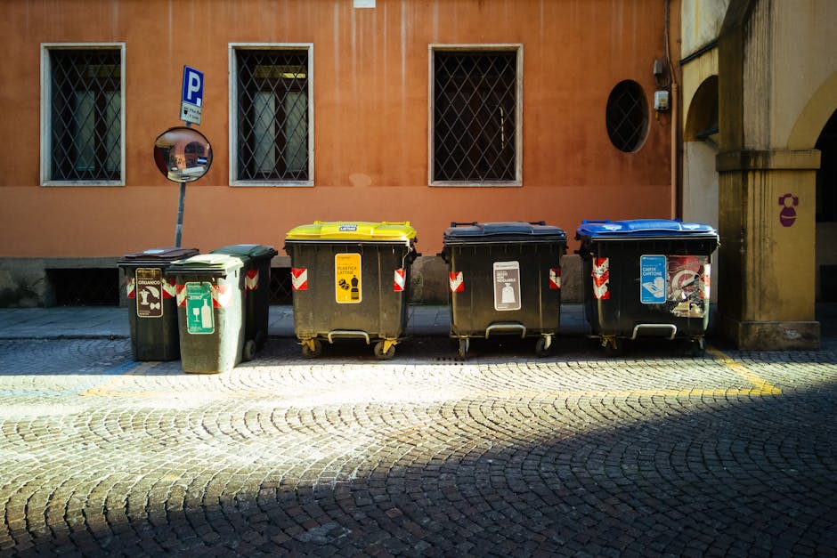 Four large outdoor waste and recycling bins positioned on a cobblestone pavement in front of an orange building with barred windows. From left to right, the bins include a green general waste bin with